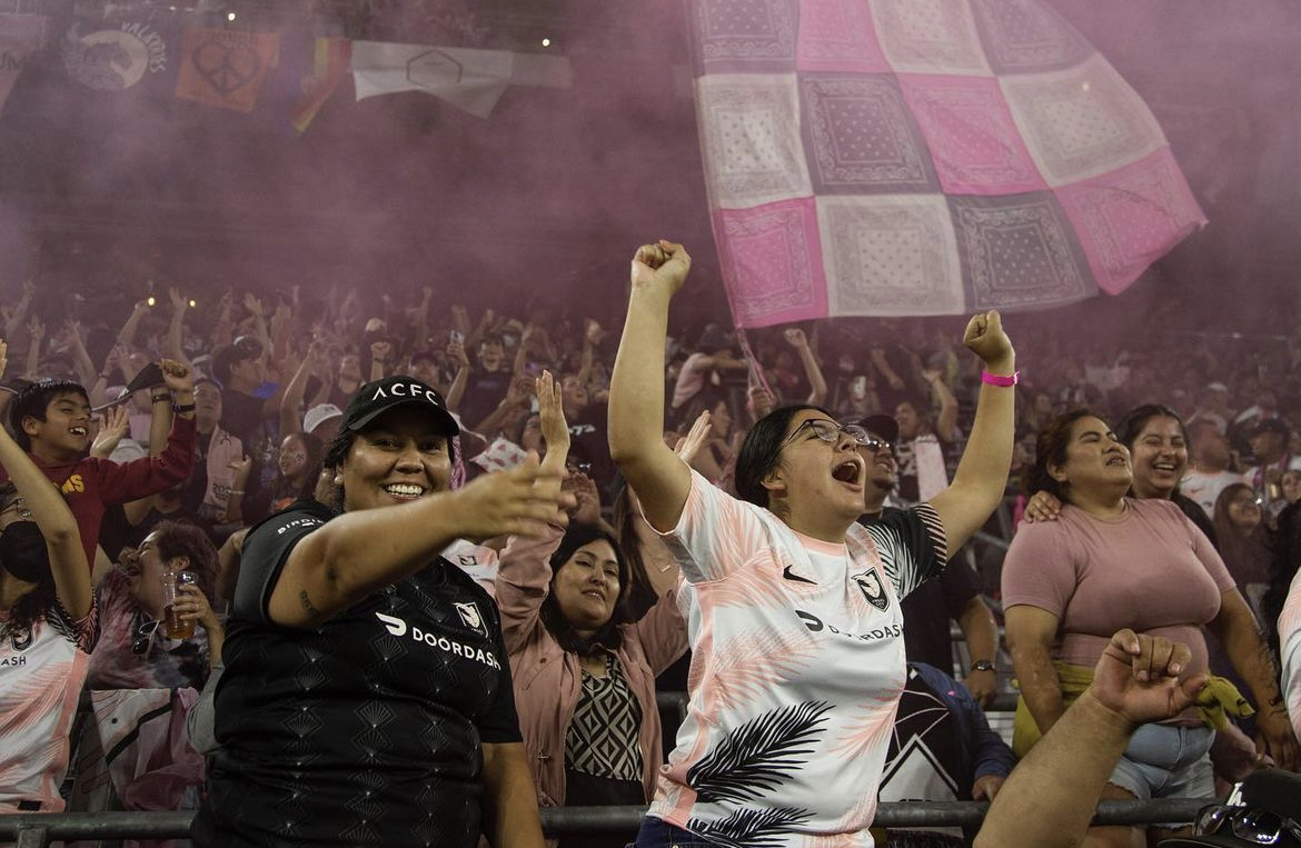 A photo of supporters cheering in La Fortaleza (supporters section) during an Angel City match, with a large flag, banners, and pink smoke in the background.