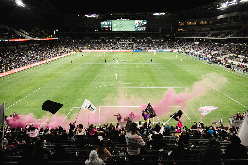 A photo from behind La Fortaleza (supporters section) during an Angel City match, with supporters, flags, and pink smoke in the foreground and the field in the background.